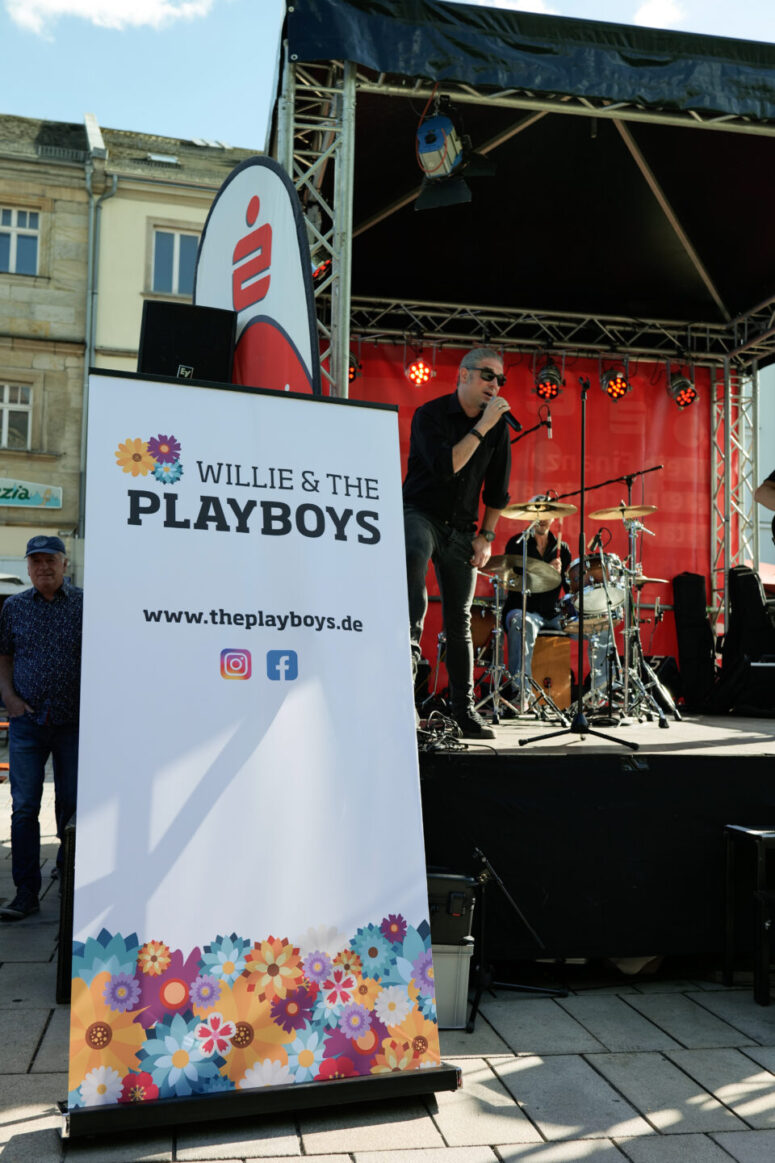 Ein Mann singt auf einer Freiluftbühne beim Bürgerfest 2026, hinter ihm sitzt ein Schlagzeuger. Im Vordergrund ist ein Schild mit der Aufschrift „Willie & The Playboys“ zu sehen, auf dem Website- und Social-Media-Symbole abgebildet sind und das mit bunten Blumen verziert ist.