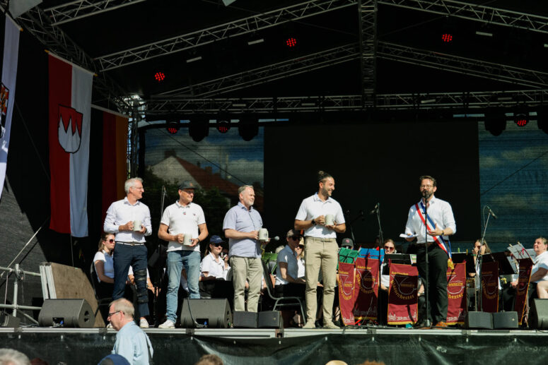 Sechs Männer stehen auf einer Freiluftbühne beim Bürgerfest 2026; einige halten Getränke in der Hand, einer spricht ins Mikrofon, während hinter ihnen Musiker spielen. Große Fahnen und Transparente rahmen die lebhafte Veranstaltung ein.