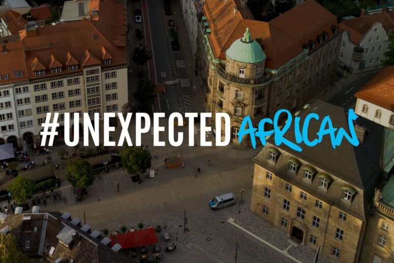 Luftaufnahme eines Stadtplatzes im europäischen Stil in Bayreuth, über das Bild ist der Text #UNEXPECTED AFRICAN in fetter Schrift geschrieben.