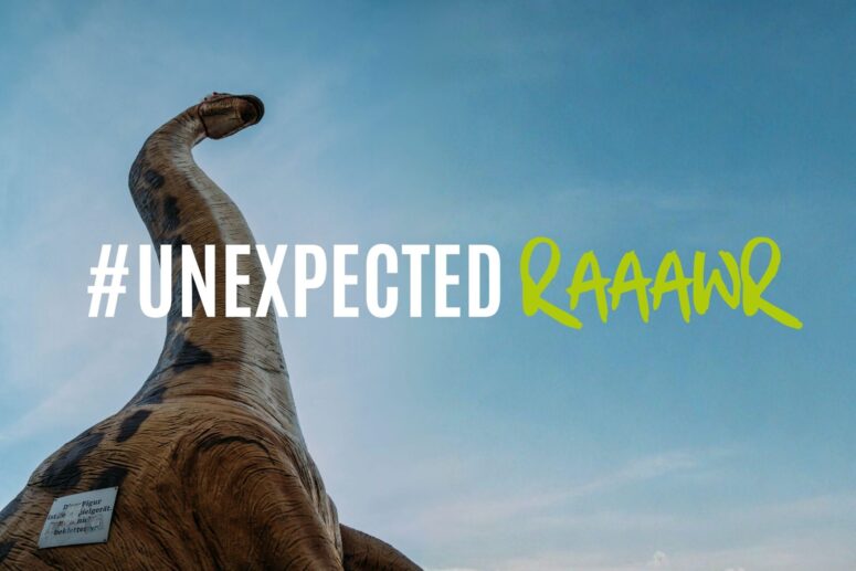 Unter dem blauen Himmel in Bayreuth steht eine große Dinosaurierstatue. Über dem Bild ist in fetter Schrift der Schriftzug #UNEXPECTED RAAAWr zu lesen, wobei „RAAAWr“ in leuchtend grüner Handschrift geschrieben ist.