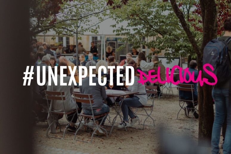 Im Herzen von Bayreuth sitzen Menschen an Tischen im Freien in einem von Bäumen gesäumten Innenhof, essen und unterhalten sich. In der Mitte ist in großen Buchstaben in Weiß und Rosa „#UNEXPECTED DELICIOUS“ zu lesen.