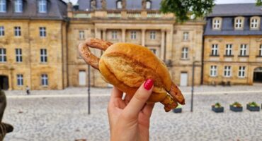 Eine Hand mit rot lackierten Fingernägeln hält vor einem historischen Steingebäude in einem gepflasterten Innenhof ein Sandwich mit einer ganzen Wurst in Brezelteigmantel und einen Genießer-Pass in der Hand.