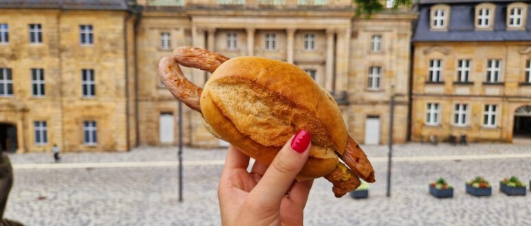 Eine Hand mit rot lackierten Fingernägeln hält vor einem historischen Steingebäude in einem gepflasterten Innenhof ein Sandwich mit einer ganzen Wurst in Brezelteigmantel und einen Genießer-Pass in der Hand.