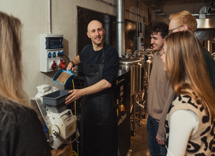 Ein Mann in einer Schürze schüttet Getreide in eine Maschine, während vier Mitglieder der „Gruppen“ in einer Brauerei oder einer Großküche zusehen und lächeln.
