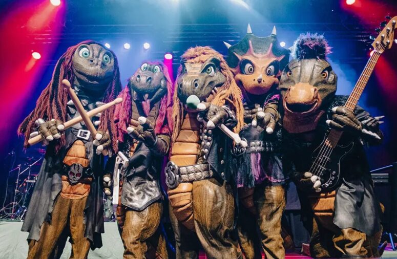 Fünf Personen in Kostümen von Dinosauriern und prähistorischen Tieren posieren auf der Bühne im Schein bunter Lichter und halten Musikinstrumente wie Trommelstöcke und eine Gitarre in den Händen.