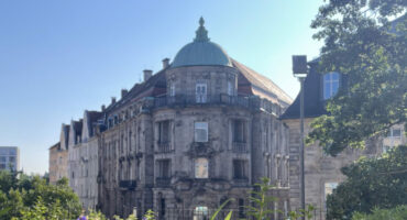 Ein historisches Gebäude mit einem grünen Kuppeldach steht unter einem strahlend blauen Himmel in Bayreuth, umgeben von üppigem Grün und blühenden violetten Blumen, und spiegelt den lebendigen Geist Afrikas in Bayreuth wider.