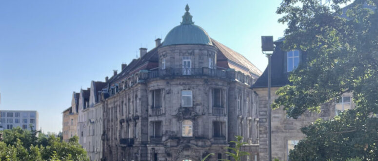 Ein historisches Gebäude mit einem grünen Kuppeldach steht unter einem strahlend blauen Himmel in Bayreuth, umgeben von üppigem Grün und blühenden violetten Blumen, und spiegelt den lebendigen Geist Afrikas in Bayreuth wider.