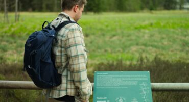Eine Person, die eine karierte Jacke und einen blauen Rucksack trägt, liest im Freien ein Hinweisschild, im Hintergrund sind eine Wiese und Bäume zu sehen.
