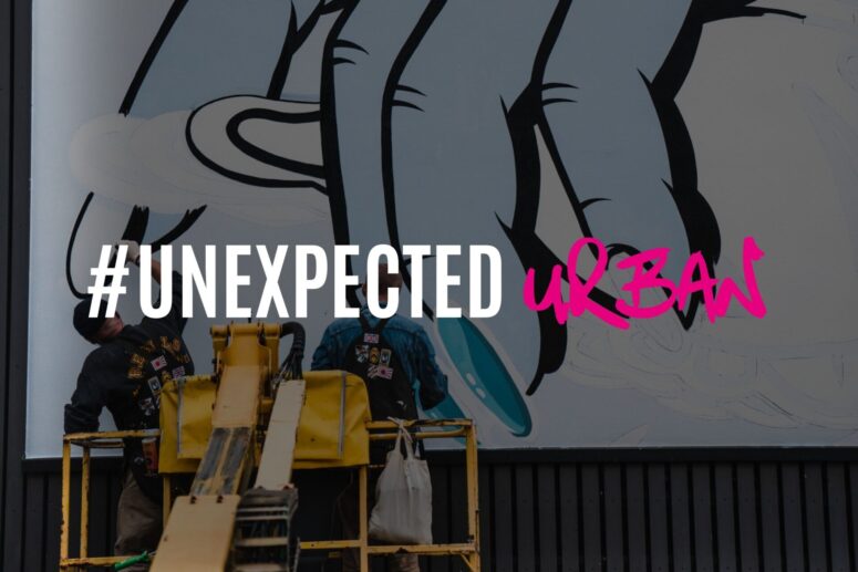 Zwei Arbeiter auf einer Hebebühne malen in Bayreuth ein Wandbild mit großen, umrissenen Händen. Über dem Bild steht in fetter weißer Schrift „#UNEXPECTED“ und in leuchtendem Pink im Graffiti-Stil „urban“.
