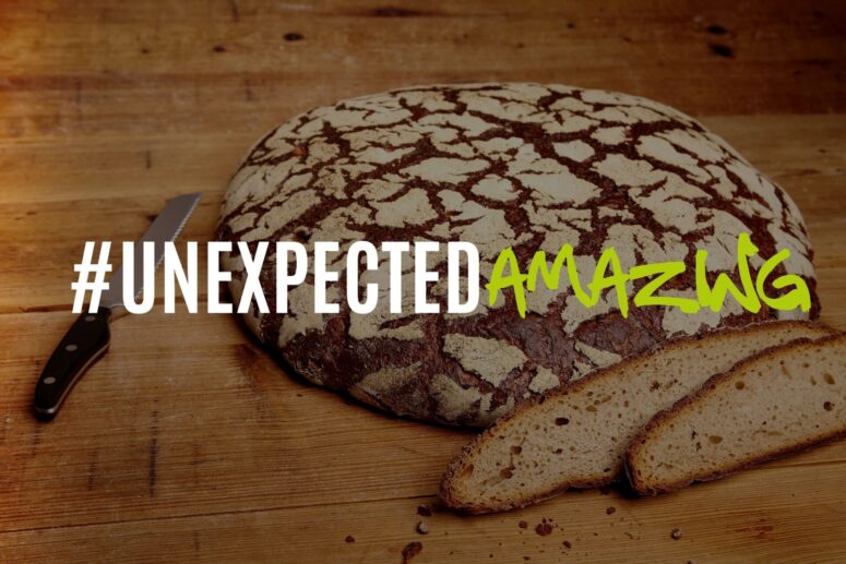 Ein rundes rustikales Brot auf einem Holztisch, von dem zwei Scheiben abgeschnitten sind; daneben liegt ein gezacktes Messer – das den handwerklichen Charme von Bayreuth vermittelt. Der Text lautet: #UNEXPECTEDAMAZING, wobei „AMAZING“ in grüner Schreibschrift geschrieben ist.