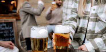 Zwei Menschen stoßen im Freien an einem Holztisch mit Biergläsern an, während im unscharfen Hintergrund weitere Freunde miteinander plaudern und an ihren Getränken nippen.