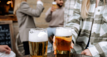 Zwei Menschen stoßen im Freien an einem Holztisch mit Biergläsern an, während im unscharfen Hintergrund weitere Freunde miteinander plaudern und an ihren Getränken nippen.