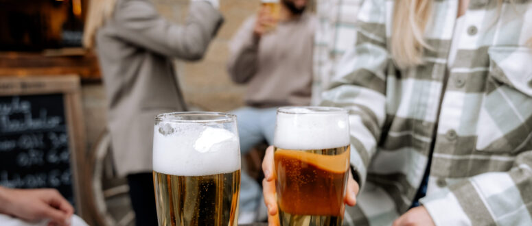 Zwei Menschen stoßen im Freien an einem Holztisch mit Biergläsern an, während im unscharfen Hintergrund weitere Freunde miteinander plaudern und an ihren Getränken nippen.