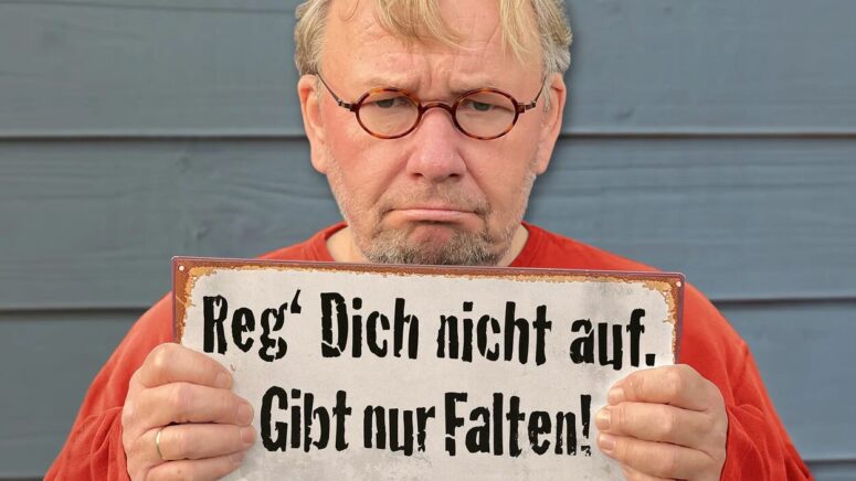 Ein älterer Mann mit runder Brille und traurigem Gesichtsausdruck hält ein Schild auf Deutsch in der Hand, auf dem steht: „Reg Dich nicht auf. Gibt nur Falten!“ Die blaue Fassade im Hintergrund lässt vermuten, dass er sich möglicherweise in der Nähe des Friedrichsforums befindet.