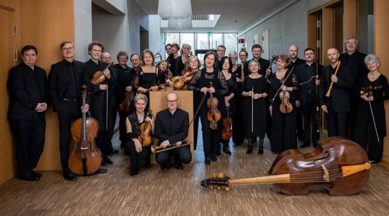 Eine Gruppe schwarz gekleideter Musiker, die Saiteninstrumente in den Händen halten, steht gemeinsam in einem Flur. Im Vordergrund liegt ein Kontrabass auf dem Boden. Die Gruppe posiert und lächelt in die Kamera.