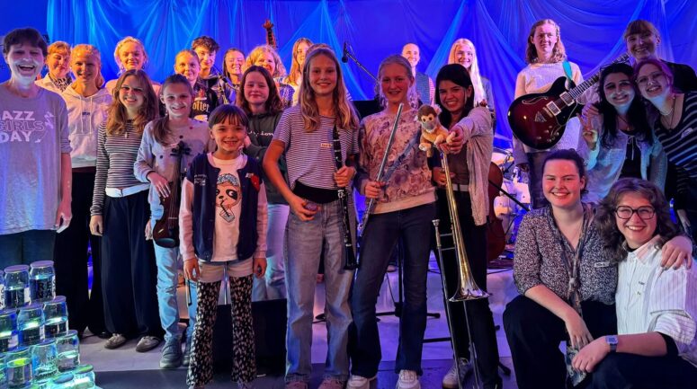 Eine große Gruppe lächelnder junger Frauen und Mädchen mit Musikinstrumenten posiert gemeinsam auf der Bühne im Friedrichsforum unter blauem Licht, was auf eine Musikveranstaltung oder ein Konzert hindeutet.