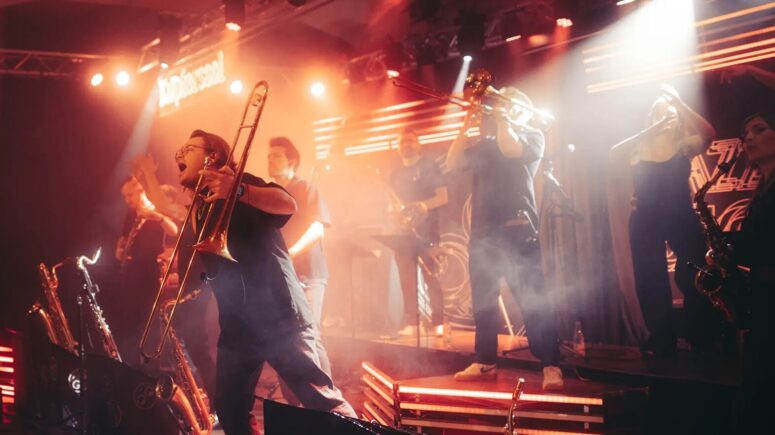 Im Friedrichsforum tritt eine lebhafte Band mit Blechblasinstrumenten unter dramatischer orangefarbener Beleuchtung auf, während ein begeisterter Musiker vorne steht, eine Posaune in der Hand hält und jubelt.