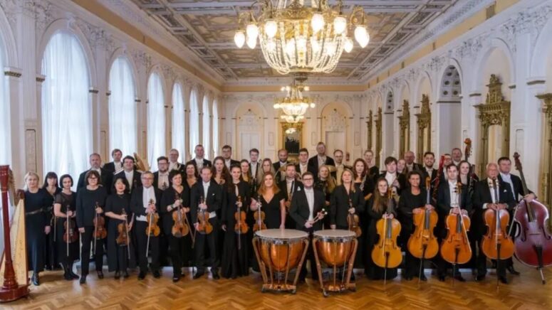 Ein großes Orchester steht in einem prächtigen, reich verzierten Saal mit Kronleuchtern und hohen Fenstern. Die Musiker halten verschiedene Instrumente in den Händen, darunter Geigen, Celli, Blechblasinstrumente, Holzblasinstrumente und Schlaginstrumente.