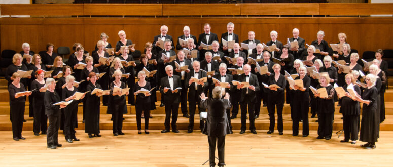 Auf der Bühne eines Konzertsaals im Friedrichsforum singt ein großer Chor aus Männern und Frauen. Vor ihnen steht ein Dirigent und leitet den Chor. Alle sind in formeller schwarzer Kleidung gekleidet und halten Notenblätter in den Händen. Im Hintergrund sind leere Sitzplätze zu sehen.