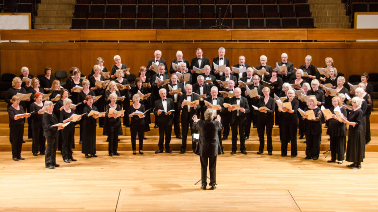 Auf der Bühne eines Konzertsaals im Friedrichsforum singt ein großer Chor aus Männern und Frauen. Vor ihnen steht ein Dirigent und leitet den Chor. Alle sind in formeller schwarzer Kleidung gekleidet und halten Notenblätter in den Händen. Im Hintergrund sind leere Sitzplätze zu sehen.