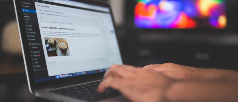 Hände, die auf der Tastatur eines Laptops tippen und einen WordPress-Blogbeitrag über eine unvergessliche Busreise bearbeiten, während auf dem Bildschirm ein Foto von einer Tasse Kaffee zu sehen ist. Im Hintergrund ist ein unscharfer, farbenfroher Monitor zu erkennen.