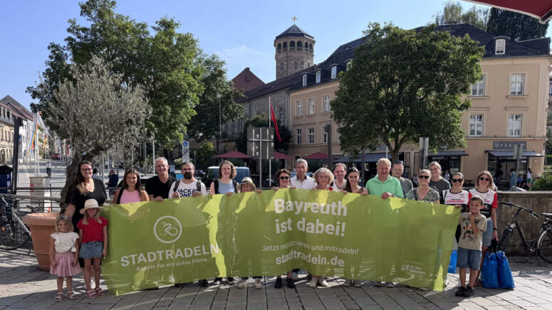 Eine Gruppe von Menschen unterschiedlichen Alters hält auf einem sonnigen Stadtplatz ein großes grünes STADTRADELN-Transparent hoch, im Hintergrund sind Bäume, Gebäude und ein Turm zu sehen.