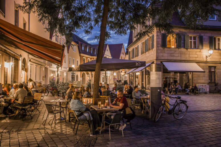 In der Abenddämmerung sitzen Menschen an den Tischen eines Straßencafés auf einer Kopfsteinpflasterstraße, während warmes Licht aus den umliegenden Gebäuden strahlt und in der Nähe Fahrräder geparkt sind. Bäume und Markisen spenden der lebhaften Szene Schatten.