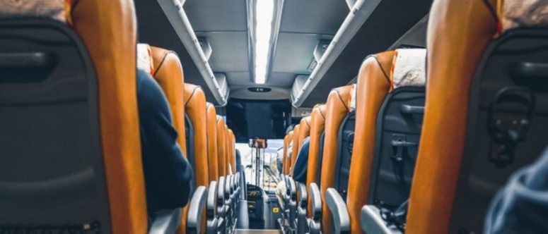Blick vom hinteren Teil eines Busses aus, den Gang entlang zwischen Reihen orangefarbener und grauer Sitze, mit Fahrgästen auf beiden Seiten und hellen Deckenleuchten – eine klassische Momentaufnahme einer Busreise in Bewegung.