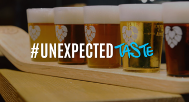 Auf einem Holzbrett stehen fünf Gläser Bier mit Schaum in einer Reihe. Der darüberliegende Text lautet „#UNEXPECTED TASTE“, wobei „TASTE“ in blauer Schreibschrift geschrieben ist.