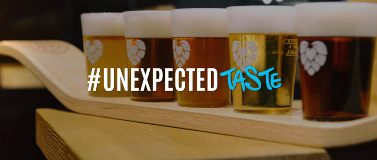 Auf einem Holzbrett stehen fünf Gläser Bier mit Schaum in einer Reihe. Der darüberliegende Text lautet „#UNEXPECTED TASTE“, wobei „TASTE“ in blauer Schreibschrift geschrieben ist.