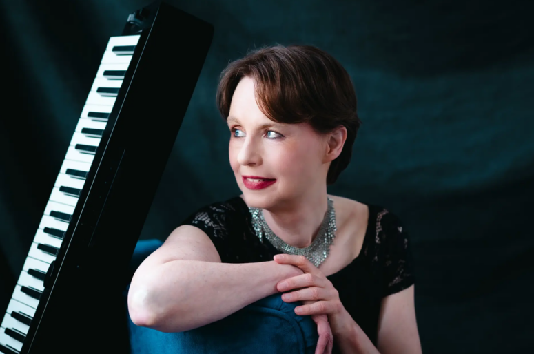 Eine Frau in einem schwarzen Kleid mit einer silbernen Halskette lehnt an einem blauen Stuhl, lächelt und blickt zur Seite; neben ihr steht vor einem dunklen Hintergrund ein elektronisches Keyboard, was die elegante Atmosphäre des Friedrichsforums heraufbeschwört.