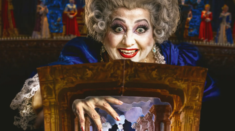 Eine Frau mit auffälligem Make-up und grauen Locken grinst breit; sie trägt ein blaues Kleid und eine Perlenkette. Sie ragt über einem kunstvoll verzierten Miniaturtheater empor, das an das Friedrichsforum erinnert, und streckt ihre Hand nach winzigen Bühnenfiguren aus.