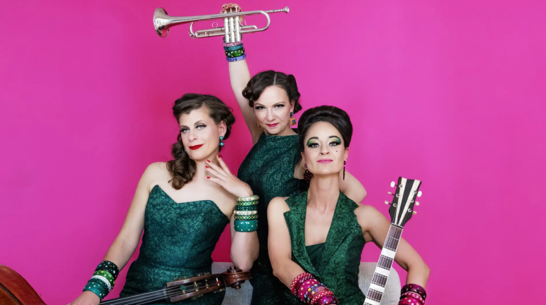 In dieser farbenfrohen Werbeanzeige für das Friedrichsforum posieren drei Frauen in grünen Kleidern vor einem leuchtend rosa Hintergrund. Eine hält ein Cello in der Hand, eine andere eine Gitarre, und die dritte hebt eine Trompete empor. Alle tragen bunte Armreifen, haben gestylte Frisuren und sind auffällig geschminkt.