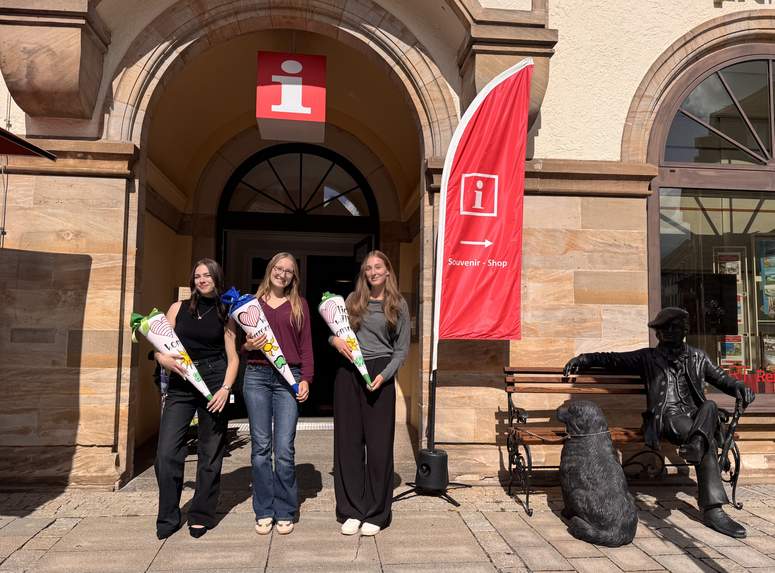 Drei lächelnde Frauen, die große, verzierte Schultüten in den Händen halten, stehen vor einem Gebäude mit einem Hinweisschild und einer roten Flagge. Auf einer Bank in der Nähe steht eine Bronzestatue eines sitzenden Mannes und eines Hundes.