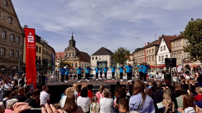 Eine Gruppe von Menschen in blauen Hemden tritt während der TOGGO-Tour auf einer Freiluftbühne auf einem Stadtplatz auf, umgeben von einem großen Publikum. Im Hintergrund sind unter einem teilweise bewölkten Himmel historische Gebäude und eine Kirche zu sehen.