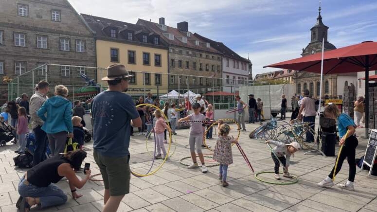 Während der TOGGO-Tour spielen Kinder und Erwachsene auf einem belebten Platz im Freien, der von historischen Gebäuden umgeben ist, mit Hula-Hoop-Reifen. Einige Leute schauen zu oder machen Fotos, während andere mitmachen. Unter einem teilweise bewölkten Himmel herrscht eine lebhafte und festliche Stimmung.