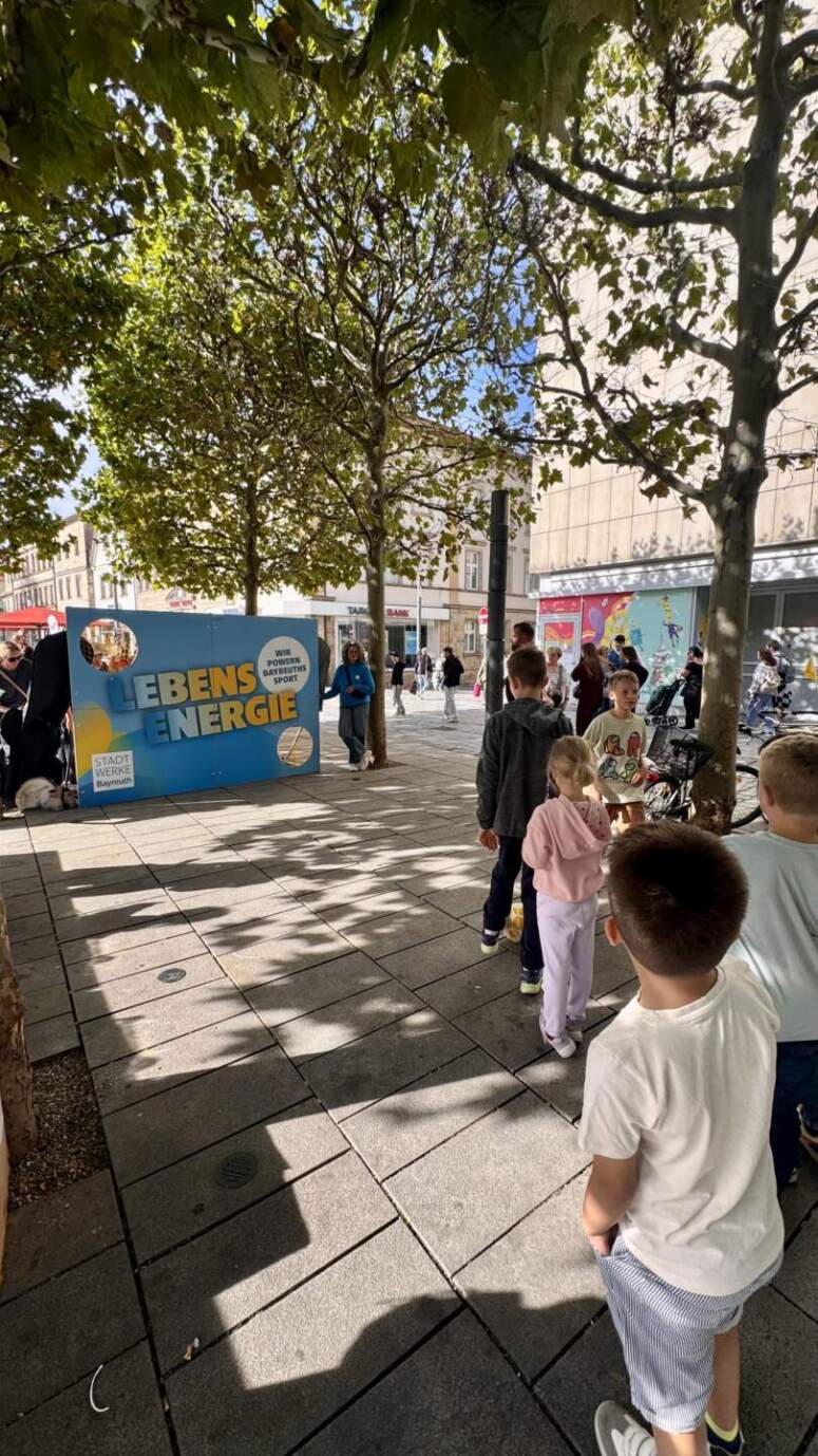 Während der lebhaften TOGGO-Tour stehen Kinder auf einem städtischen Bürgersteig in der Nähe eines blauen Ausstellungsstandes mit der Aufschrift „Lebens Energie“ in einer Reihe. Bäume spenden Schatten, und im Hintergrund sind an einem sonnigen Tag Menschen zu sehen.