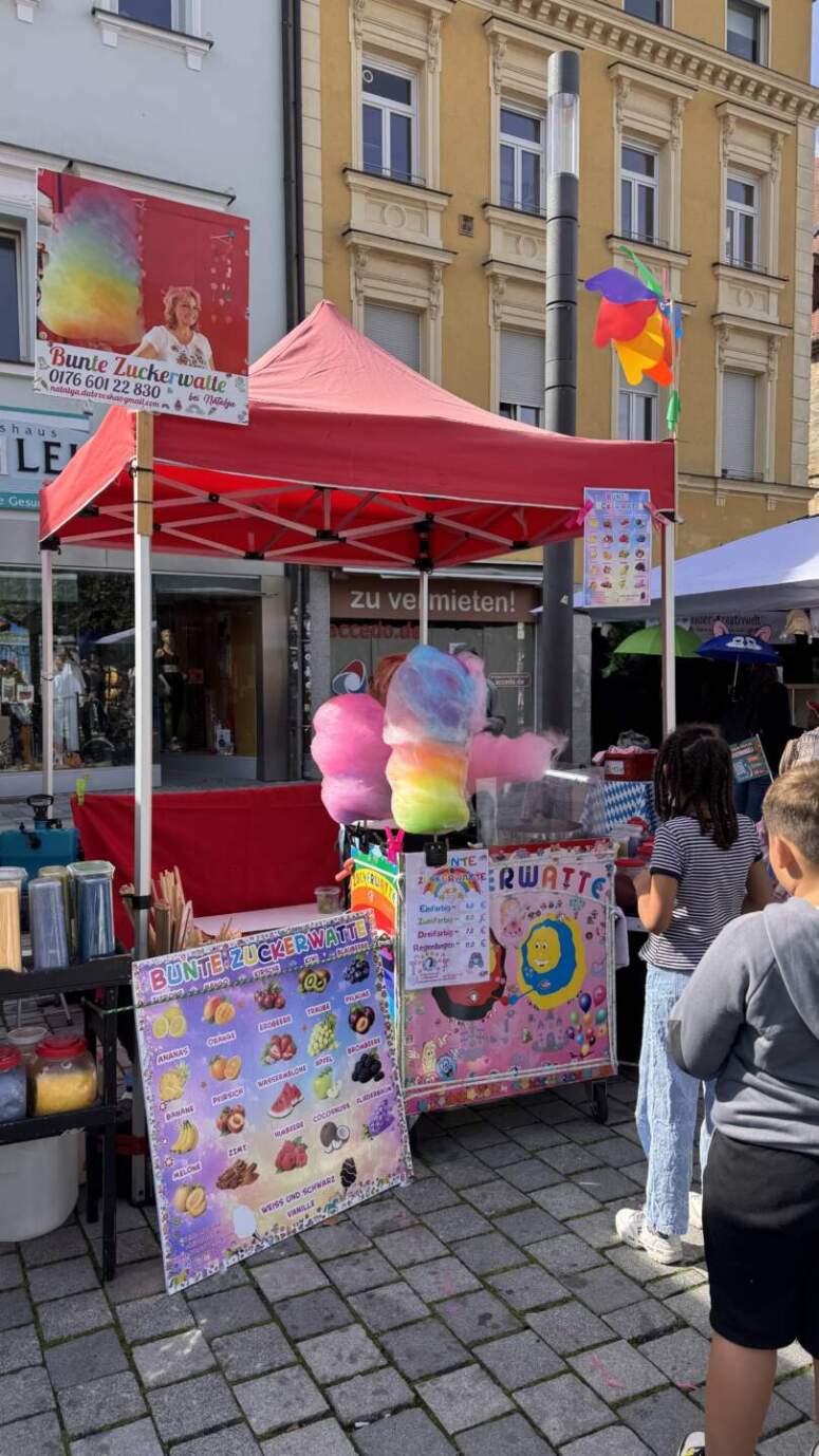 Ein farbenfroher Zuckerwatte-Stand mit rotem Vordach, leuchtenden Zuckerwatte-Auslagen und Speisekarten fällt bei der lebhaften TOGGO-Tour-Veranstaltung besonders ins Auge. Vor dem Stand versammeln sich mehrere Kinder, die aufgeregt ihre Leckereien aussuchen, während im Hintergrund Gebäude zu sehen sind.