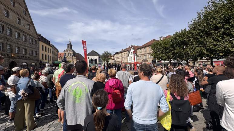 Auf einem Stadtplatz versammelt sich draußen eine große Menschenmenge vor einer Bühne mit roten Bannern und „TOGGO-Tour“-Schildern. Es ist sonnig, und der Platz ist von historischen Gebäuden umgeben. Menschen jeden Alters sind anwesend; einige machen Fotos oder schauen aufmerksam zu.