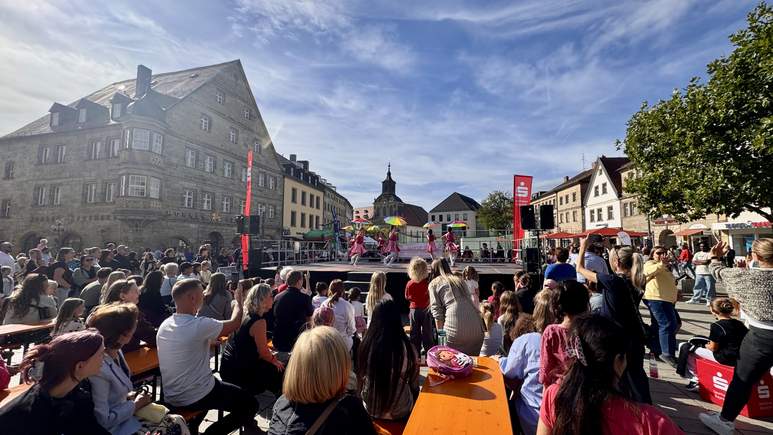 Eine Menschenmenge beobachtet Tänzer, die während der TOGGO-Tour auf einer Freiluftbühne auf einem von historischen Gebäuden umgebenen Stadtplatz unter blauem Himmel mit vereinzelten Wolken auftreten.