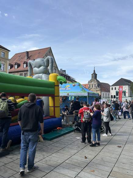 Unter blauem Himmel versammeln sich Menschen bei einer Veranstaltung im Freien, die Teil der lebhaften TOGGO-Tour ist – mit einer Hüpfburg in Form eines Elefanten, bunten Zelten und historischen Gebäuden im Hintergrund.