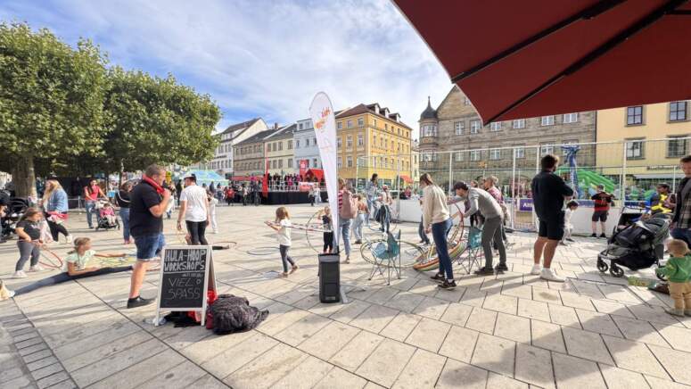 Auf einem sonnigen Platz versammeln sich Menschen im Freien: Kinder spielen, Erwachsene schauen zu, es gibt Banner und bunte Dekorationen. Im Hintergrund reihen sich Gebäude aneinander, und ein Schild wirbt für Workshops. Einige Leute hören einem Redner am Mikrofon zu – als Teil der lebhaften TOGGO-Tour-Veranstaltung.