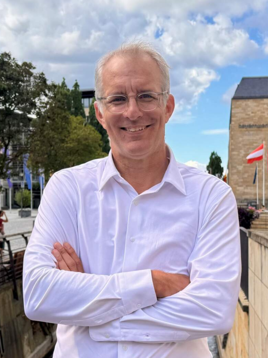Ein Mann mit kurzen grauen Haaren und Brille lächelt mit verschränkten Armen; er trägt ein weißes Hemd und steht im Freien in der Nähe von Gebäuden und Bäumen unter einem teilweise bewölkten Himmel und verkörpert Professionalität im Tourismusmanagement.