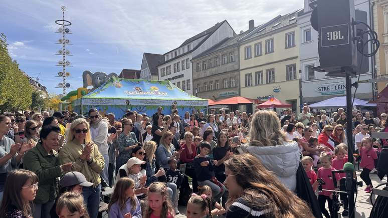 Eine große Menschenmenge, darunter viele Kinder und Erwachsene, versammelt sich im Freien bei einem lebhaften TOGGO-Tour-Straßenfest mit Zelten, bunten Ständen und einer Bühne unter strahlendem Sonnenschein.