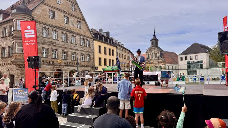 Ein Künstler mit Hut unterhält eine Menge aus Kindern und Erwachsenen auf einer Freiluftbühne auf einem historischen europäischen Platz, umgeben von alten Gebäuden und roten Fahnen, im Rahmen der lebhaften TOGGO-Tour.
