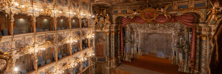 Ein prächtiges, kunstvoll verziertes Theater mit vergoldeten Balkonen, bemalter Decke, aufwendigen Wandmalereien und einer großen Bühne mit roten Vorhängen, gesehen von einer Ecke im Obergeschoss. Unten im Zuschauerraum reihen sich die Sitzreihen aneinander.