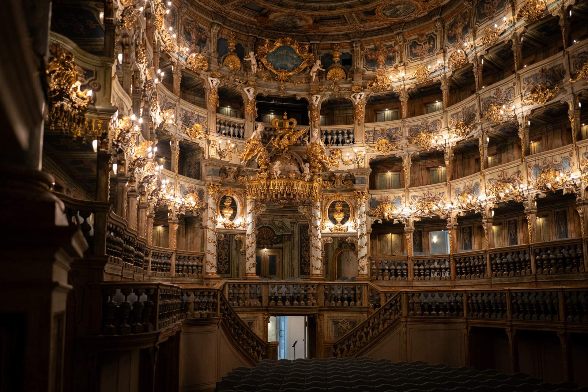 Ein prächtiges, kunstvoll gestaltetes Theaterinterieur mit goldenen Verzierungen, Balkonen, Kronleuchtern und detailreichen Kunstwerken, beleuchtet von warmem Licht; im unteren Vordergrund sind leere Sitze zu sehen.