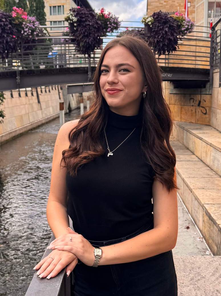 Eine junge Frau mit langen braunen Haaren und einem schwarzen, ärmellosen Oberteil steht an einem Geländer neben einem Kanal und lächelt in die Kamera. Blumen in Pflanzkübeln hängen von der Brücke über dem Wasser herab.