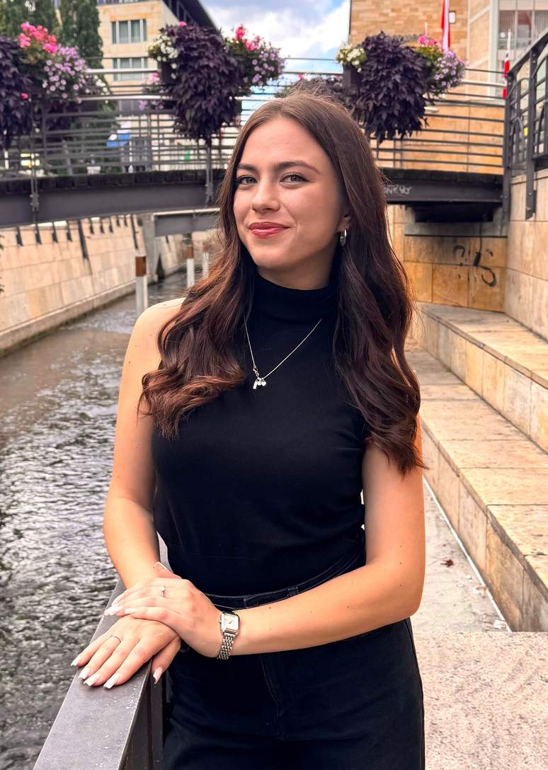 Eine junge Frau mit langen braunen Haaren, die ein schwarzes, ärmelloses Oberteil trägt, steht an einem Kanal mit Steinmauern und lächelt in die Kamera. Im Hintergrund sind Blumen in Pflanzkübeln und eine Brücke zu sehen.