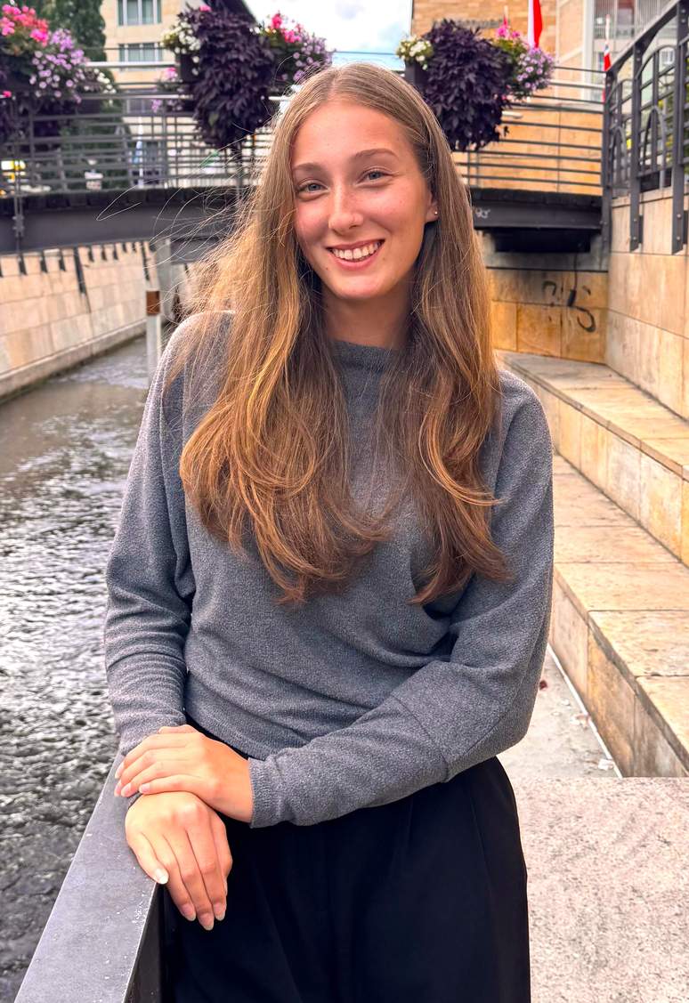 Eine junge Frau mit langen braunen Haaren, die einen grauen Pullover trägt, lächelt, während sie an einem Kanal steht, im Hintergrund sind Geländer, Steinmauern und Topfpflanzen zu sehen.